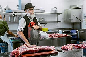 Photo: Sustine, Rowan Thornhill Un homme se tient devant un plan de travail en acier inoxydable avec un gros morceau de viande.